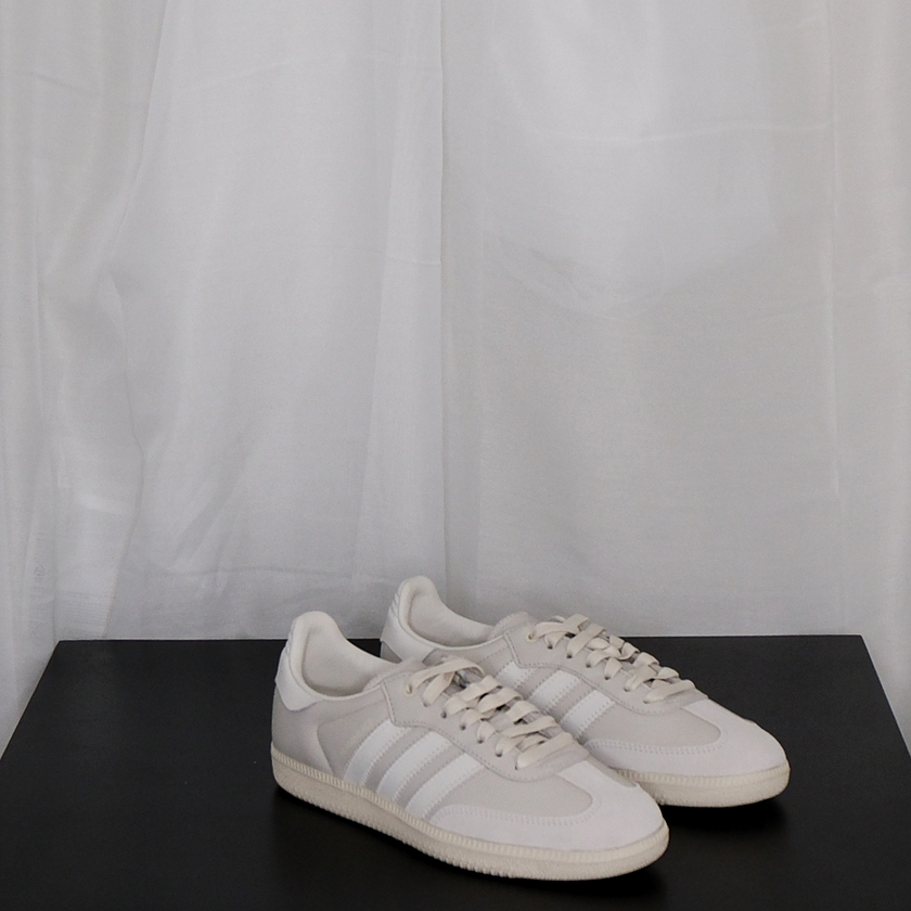 Pair of beige Adidas sneakers on a black surface with a white curtain background